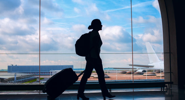 Eine Frau geht mit einem Rollkoffer in einem Flughafen an einem großen Fenster vorbei. Im Hintergrund sind parkende Flugzeuge zu sehen.