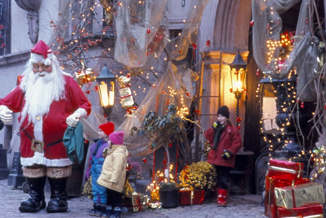 Weihnachtsschmuck und Weihnachtsgeschenke schmücken den Hintergrund vom Weihnachtsmann der in einer Straße von Riquewihr (Elsass) steht.