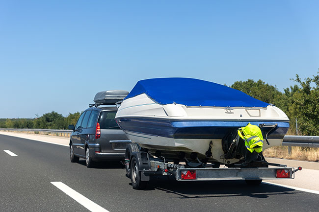 Ein Familien-Van zieht einen überdachten Bootsanhänger und fährt auf einer leeren Autobahn unter strahlend blauem Himmel, umgeben von Hügeln. Der strahlende Sommertag vermittelt ein Gefühl von Freiheit, Reiseabenteuer und mobilem Lebensstil auf einer malerischen Strecke