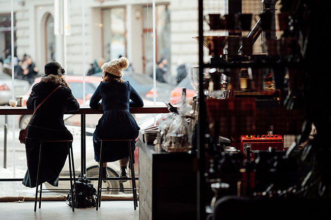 Zwei Frauen sitzen an einem Hochtisch in einem Kaffee in Paris an einer großen Fensterfront und unterhalten sich