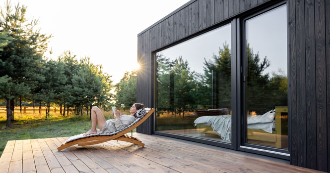 Junge Frau, die auf einer Sonnenliege ruht und auf einem Liege auf einer Holzterrasse in der Nähe eines modernen Blockhauses mit Panoramafenstern in der Nähe eines Kiefernwaldes und liest.